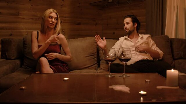 A blonde in a burgundy dress and a brunette man in an unbuttoned shirt continue a calmer dispute on a beige sofa. Occasional smiles hint tension fading, while soft candlelight sets a mellow tone.
