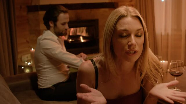 A blonde in black lingerie speaks rapidly with wide eyes and tense gestures, bordering on hysteria. Calm brunette man in white shirt sits by the fireplace, quietly observing under flickering candles.