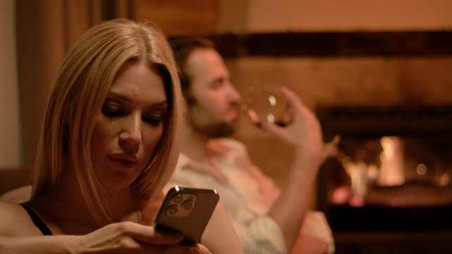 In the foreground, a blonde in black lingerie scrolls intently on her smartphone. Behind her, a brunette man in a white shirt casually sips wine by a glowing fireplace and soft candlelight.