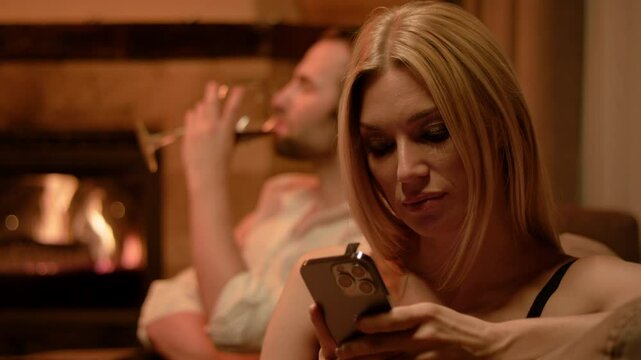 A blonde in black lingerie calmly checks her smartphone, face relaxed as her fingers glide across the screen. In the background, a brunette man in white shirt leisurely drinks wine near the fireplace