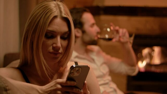 A blonde in black lingerie studies something on her smartphone with quiet focus. Behind her, a brunette man in a white shirt takes a small sip of wine. The fireplace glows with gentle light.
