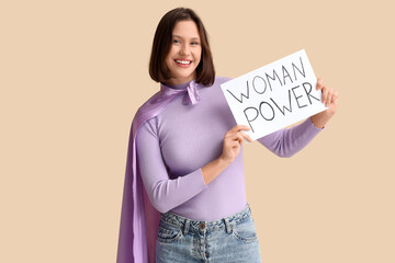 Beautiful young woman with super hero cape holding WOMAN POWER sign on beige background. Women's...