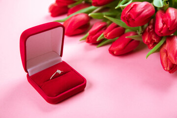 Diamond ring in box lais on a big bouquet of red tulips on pink background. Symbol of love. The soul union. Jewelry gift certificate. Holiday business card. Engagement concept. Copy space. Side view