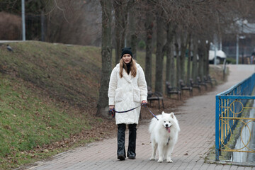 A person dressed warmly strolls through a park, holding the leash of a fluffy white samoyed. The surroundings are tranquil with benches and trees