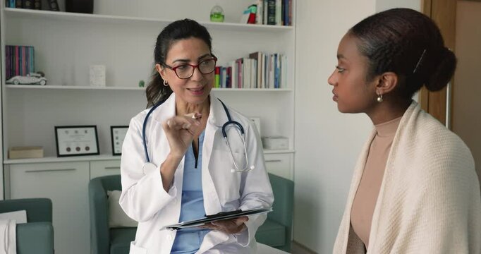 Smiling middle-aged female doctor wear white coat holds clipboard, asks about health complaints and disease symptoms, talking to young African woman patient during her visit at hospital. Healthcare