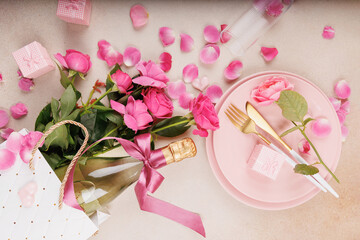 A cozy and elegant table with fresh roses, a champagne bottle with a bow, golden cutlery, and soft pink decorations.