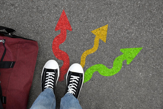 Man standing near colorful arrows pointing in different directions on asphalt road, closeup. Concepts of choice, decision, alternative way