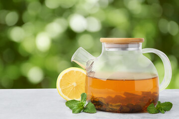 Aromatic mint tea in glass teapot, fresh leaves and lemon on light grey table outdoors. Space for text