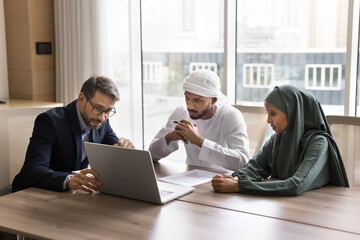 European financial advisor consults Arabian couple using laptop, talk about contract details, showing presentation, explain conditions met in office. Sell and buy services, commercial deal discussion