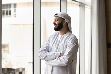 Arab businessman in robe stands by window, gazing out with thoughtful expression reflecting on achievements, strategizing about future business ventures, contemplating personal growth, upcoming goals