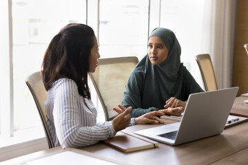 Multicultural businesswomen having conversation seated at table with laptop. Arabian female dressed in hijab talks to colleagues solve work-related matters, discuss project details, work on joint task