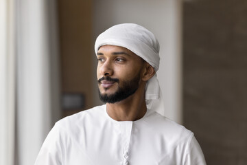 Confident Middle Eastern man stands indoor, gazing thoughtfully into distance. Young Arabian businessman, entrepreneur dressed in traditional white dishdasha, looking aside with pensive expression