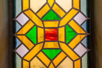 A close-up shot of a stained glass window showcasing a geometric pattern with a central red square, surrounded by yellow and green sections, against a textured background.