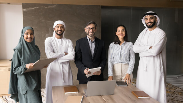 Five multicultural teammates, European and Arabian staff posing in office with positive expression, showing unity, synergy, feel satisfied with career, promotion and successful, productive teamwork