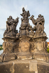 A stone bridge adorned with statues, including a prominent group featuring a crowned female figure with other figures below, overlooking a river and cityscape under a partly cloudy sky.
