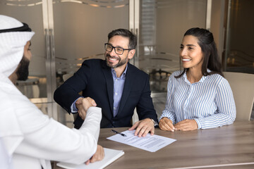 Fototapeta premium Manager and clients sealing professional agreement with handshake. Happy European spouses signing contract, shaking hands with Arabian male professional, express gratitude for getting services. Deal