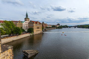Obraz premium A wide river with boats sailing under a cloudy sky, bordered by stone embankments and historic buildings with red-tiled roofs, featuring a prominent tower with a clock