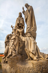 Obraz premium A statue group featuring several figures with detailed clothing, positioned atop a stone pedestal against a backdrop of a cityscape with red-roofed buildings and greenery under a cloudy sky.