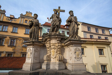 A statue group featuring three figures with detailed clothing, including one holding a cross, positioned against a backdrop of historic buildings with colorful facades and a red-tiled roof.