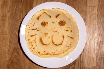 The image shows a delightful pancake cooked on the white plate, featuring a playful face with eyes, a nose, and a wide, smiling mouth. Kids meal cooking.