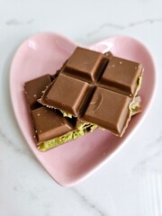 Pieces of chocolate candy bar with pistachio filling, served on a pink, heart-shaped plate against a marble background. Aesthetic Dubai chocolate.