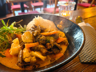 Delicious Thai curry with rice, vegetables, and fresh greens served in a rustic black bowl on a wooden table, perfect for a cozy dining experience.