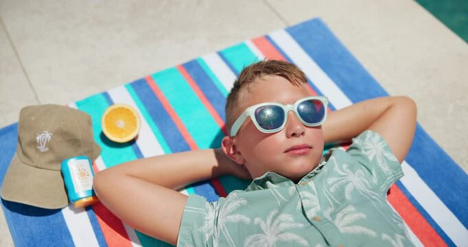 Kid, relax and sunglasses by pool outdoor with sunscreen protection, healthy fruit and cap for safety. Child, rest and eyewear for summer holiday, spf and uv treatment with vitamin c for tanning risk