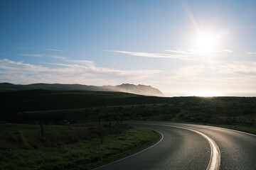 Winding coastal highway through rolling green hills at sunset near Point Reyes, California. Golden light reflecting on the asphalt, mist rising from the Pacific Ocean, dramatic, scenic road trip.