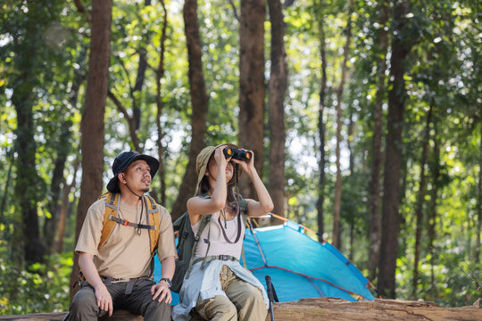 A couple of hikers sit on a fallen log in a lush forest, using binoculars to observe wildlife. Their tent is set up nearby, indicating a camping adventure. They are enjoying nature and exploration.