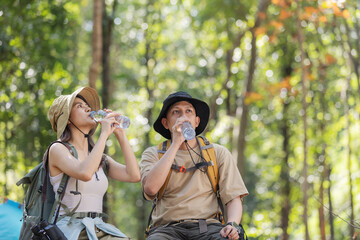 A couple of hikers take a break in the forest, drinking water from bottles to stay hydrated. They...