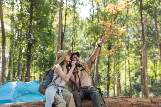 A couple of hikers sits on a fallen tree trunk in a dense forest, using binoculars for birdwatching. The man points towards something interesting while a blue tent is visible in the background
