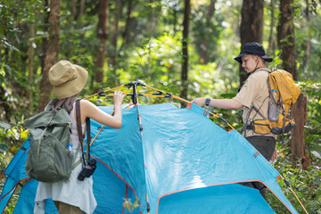 A couple sets up a blue tent in the middle of a dense forest, preparing for a camping adventure.