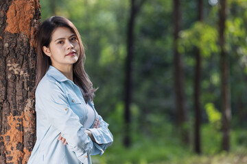 A young woman leans against a tree in a lush green forest, lost in thought. Dressed in a casual denim shirt, she gazes into the distance, reflecting on nature's beauty and the peaceful surroundings