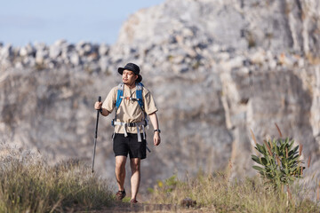 A male hiker with a backpack and trekking pole pauses on a rugged mountain trail, shielding his...