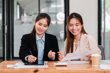 Two women engaged in a collaborative business meeting, discussing documents and using a digital tablet in a modern office environment.