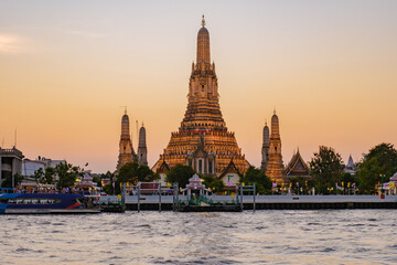 Fototapeta premium Wat Arun stands gloriously illuminated during sunset in Bangkok, casting reflections on the Chao Phraya River. Boats gently glide by as the sky transforms with warm hues.