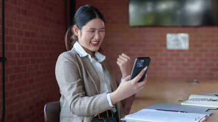 A joyful businesswoman in a beige blazer cheers while looking at her smartphone, celebrating success in a modern office setting.