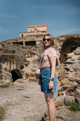 Fototapeta premium A woman in sunglasses and casual summer outfit explores the ancient rock-hewn city of Uplistsikhe, Georgia, standing against the backdrop of caves and a historic church under a bright blue sky