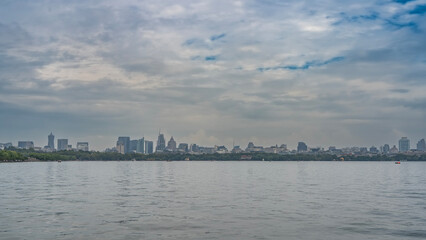 Obraz premium Beautiful calm lake. Ripples on the surface of the water. In the distance, there is green vegetation on the shore. Tall city buildings, skyscrapers against a blue sky and clouds. China. Hangzhou. 