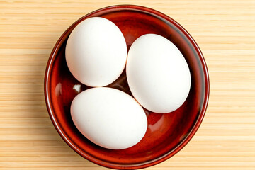 A high-quality photograph of a white ceramic bowl filled with fresh brown eggs, placed on a rustic wooden table. The background features a modern kitchen with white cabinets and a wooden countertop, 