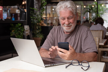 Obraz premium Attractive senior bearded man sitting in a cafe using laptop and mobile phone while enjoying a break. Modern and smiling elderly man working in a relaxed atmosphere