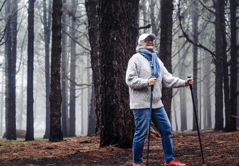 Smiling Active mature woman walking in mountain forest on a winter foggy day with the help of poles...