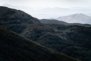 Dark, rocky hillsides descend towards a distant, hazy body of water. Layers of mountains stretch...