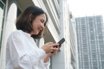 Fototapeta premium Young woman in a white shirt using smartphone outside an office building, urban scene with modern architecture in the background, smiling and engaged.