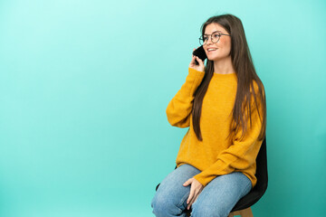 Young caucasian woman sitting on a chair isolated on blue background keeping a conversation with...