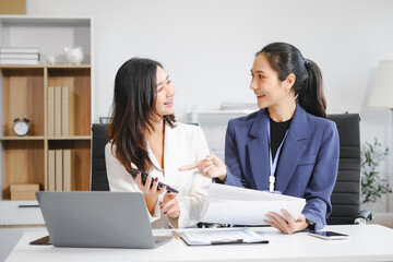 Obraz premium Two Asian female business woman sit desk,discussing business investments.They analyze financial data, explore market opportunities, and strategic growth to maximize returns ensure business success