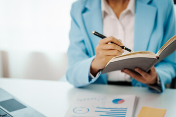An Asian female accountant works happily at her desk in the office, using a laptop to analyze financial charts. She manages business finance and online transactions for company success