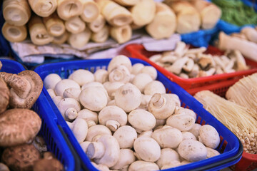 A blue basket full of white mushrooms sits on a table next to other baskets of vegetables. The mushrooms are the main focus of the image, and they appear to be fresh and ready to be eaten