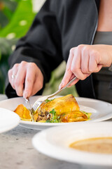 A close-up of hands using a fork and knife to cut into a delicious meal on a white plate. The dish features a golden-brown item, garnished with fresh herbs. A stylish dining setting is visible.