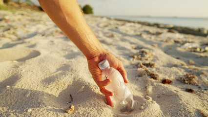 Hand collecting plastic bottle from the sand on polluted beach during golden hour. Concept of conservation, sustainability, and human pollution